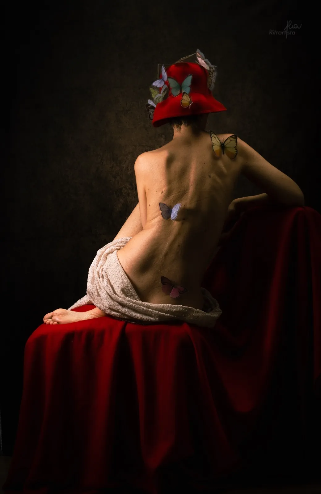 Donna di spalle con cappello rosso e farfalle, seduta su drappo rosso